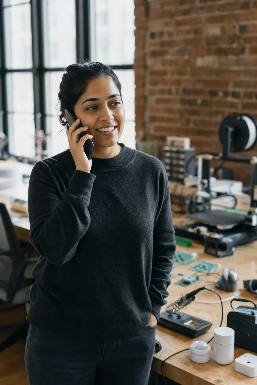 A startup founder on a phone call in a small hardware workshop, circuit boards and sensor prototypes on the bench behind her
